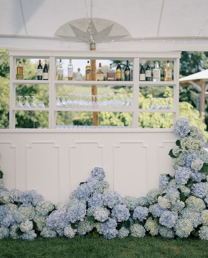 An elegant outdoor garden party bar setup with cascading light blue hydrangeas spilling across the front, white paneled bar backdrop, hanging wine glasses, lush green garden in the background, soft natural light, airy and romantic atmosphere, high-end event styling